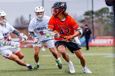 Alex Ashley, wearing an orange and maroon lacrosse uniform as he plays for the Virginia Tech Men's Lacrosse team, runs with a lacrosse stick. He is in possession of the ball. To his left, two Liberty University players, wearing white and red uniforms, try to get the ball from him.